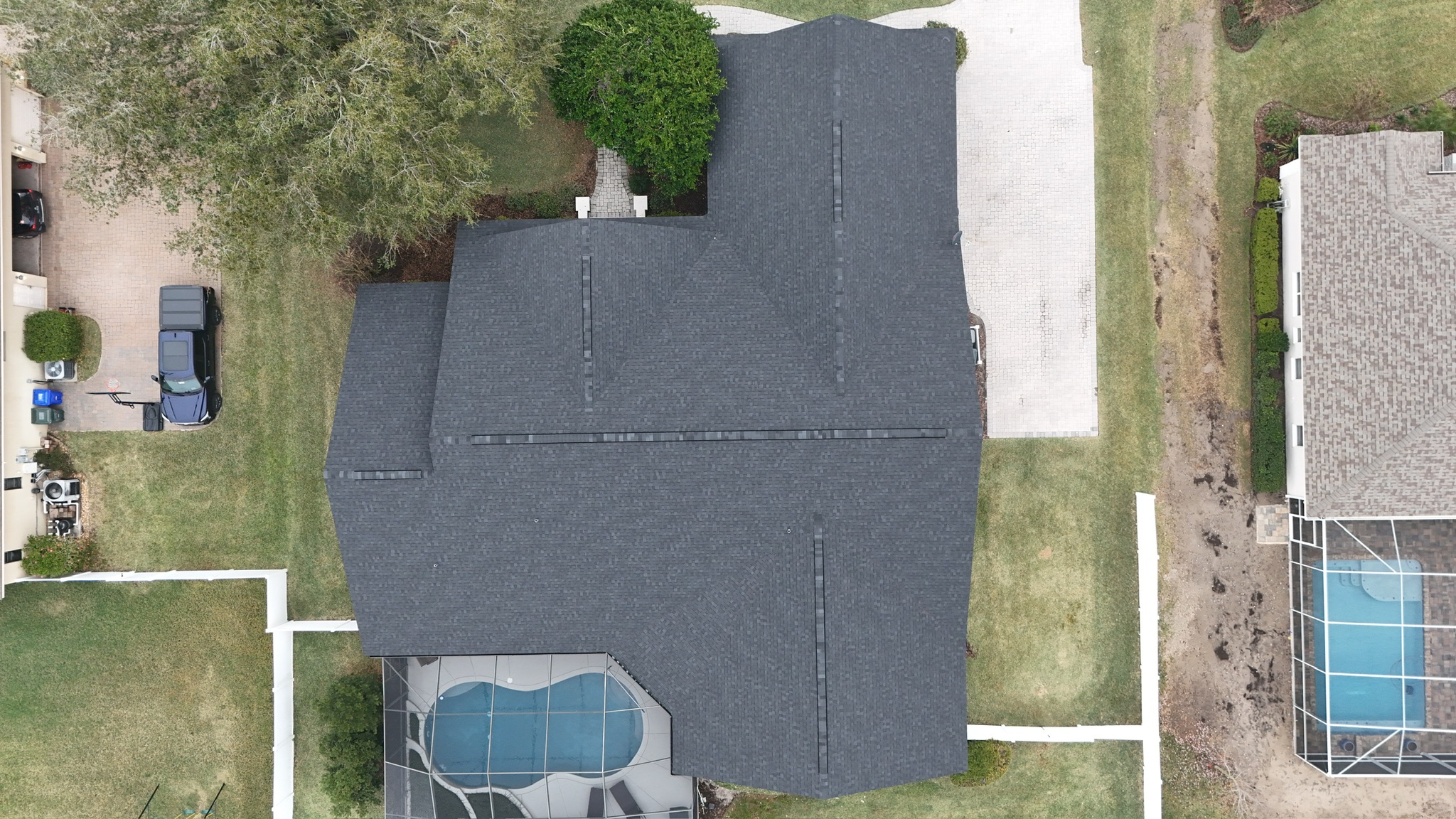Aerial view of a house with a dark gray roof, a driveway, green lawn, and a screened-in pool area at the bottom. Neighboring houses and landscaped yards are visible on either side.