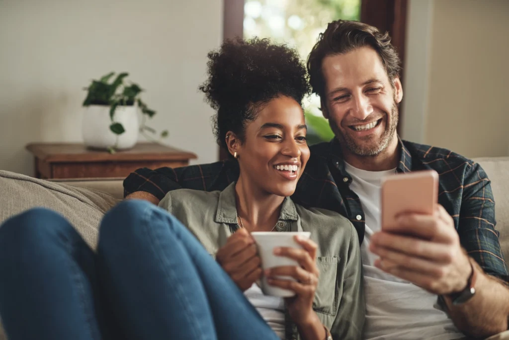 A smiling couple sits on a couch together, enjoying a cozy moment at home in Port Charlotte. The woman holds a mug and the man shows her something on his smartphone—perhaps browsing roofing Port Charlotte services. A plant is in the background.