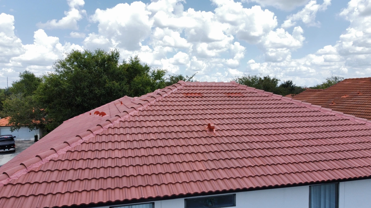 A house with a red tile roof is shown under a blue sky with fluffy white clouds. Trees and parts of nearby roofs are visible in the background, reflecting the quality craftsmanship of roofing Port Charlotte is known for.