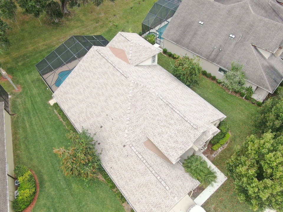 Aerial view of a beige-roofed house with a well-kept lawn, trees, and a screened pool enclosure in the backyard—showcasing expert roofing Riverview style—bordered by neighboring homes and greenery.