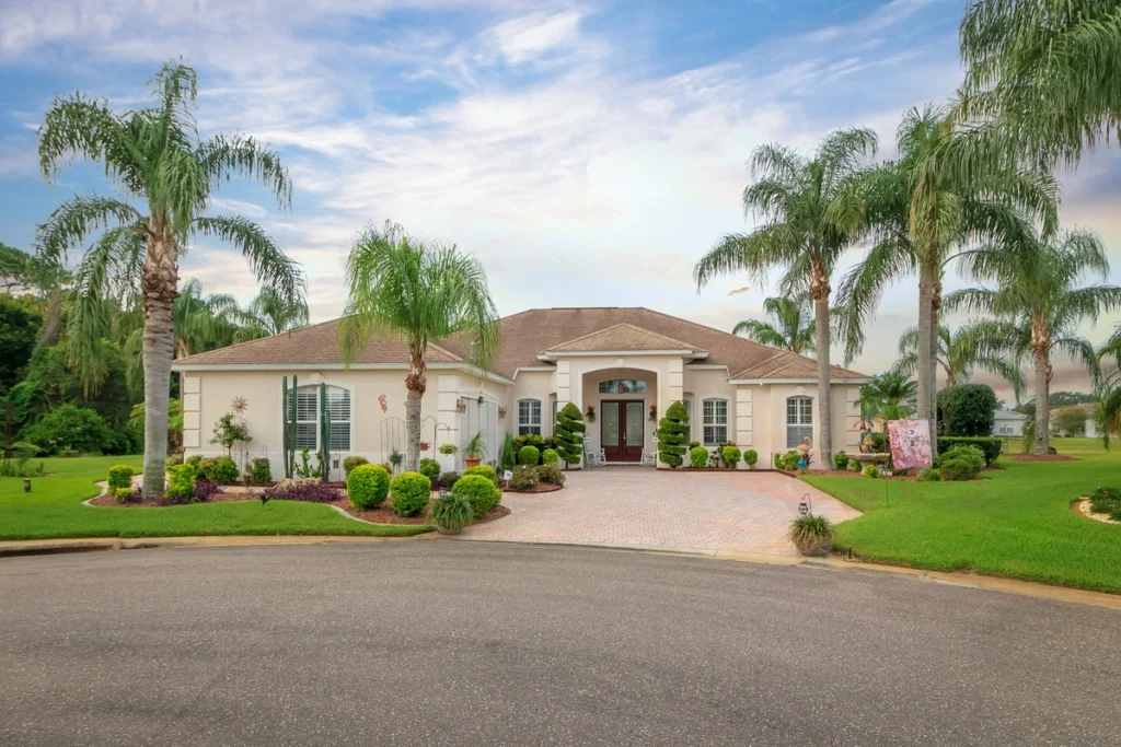 A large, single-story house with a tan exterior, palm trees, manicured lawns, and shrubs in front. The home has a paved driveway and is situated on a cul-de-sac under a partly cloudy sky.