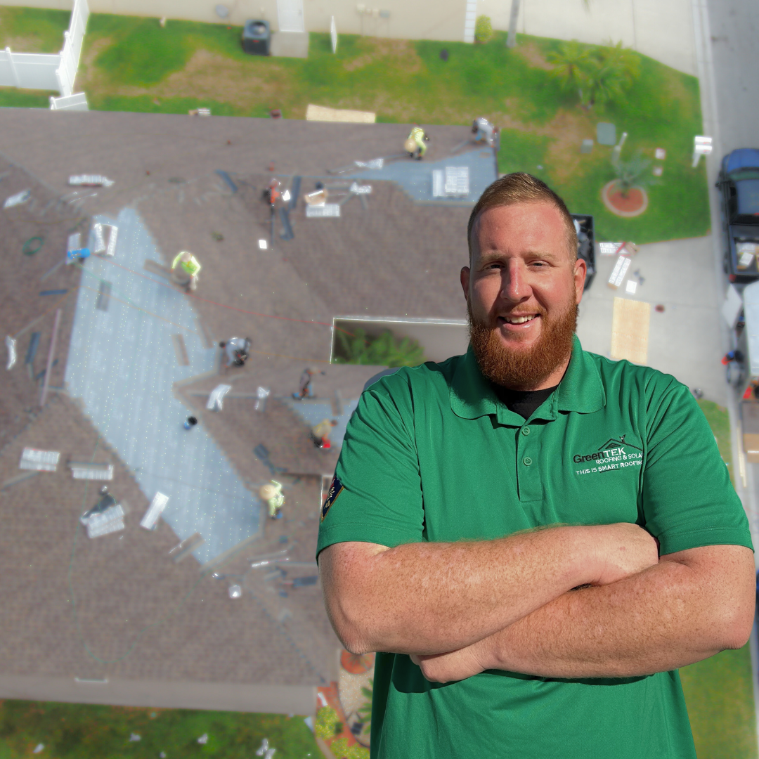 A smiling man in a green shirt stands with arms crossed in the foreground; behind him, workers repair a residential roof viewed from above.