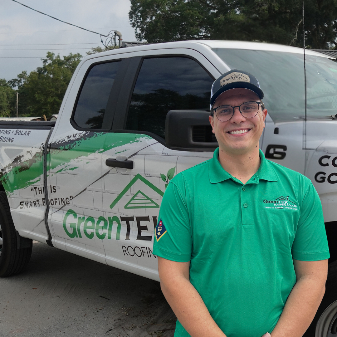 A smiling man in a green GreenTek Roofing polo and cap stands in front of a branded GreenTek Roofing truck with a white and green logo and graphics. Trees and cloudy sky are visible in the background.