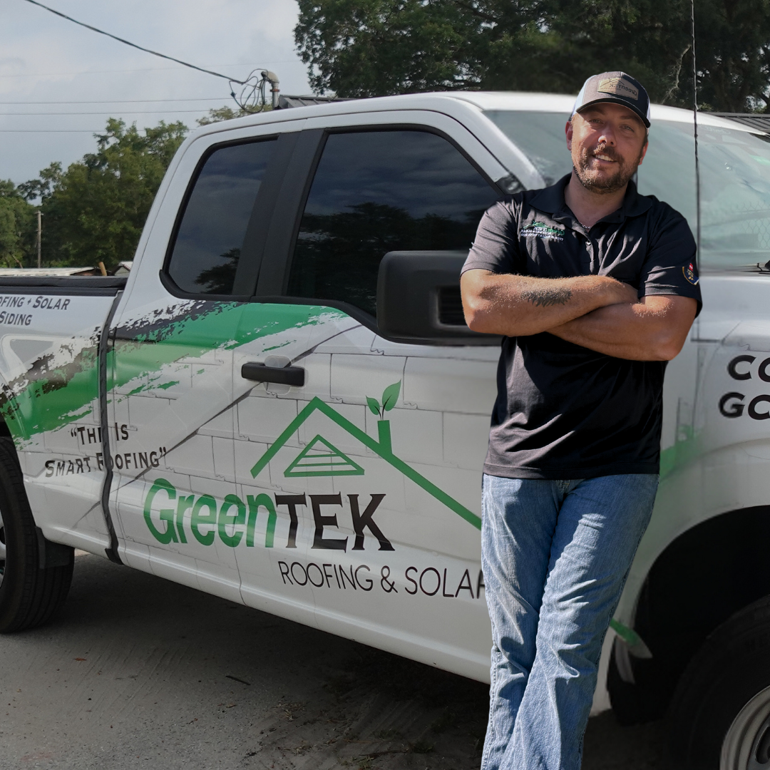 A man in a black polo shirt and cap stands smiling with arms crossed beside a white truck displaying the GreenTEK Roofing & Solar logo and slogan on the side. Trees and power lines are in the background.
