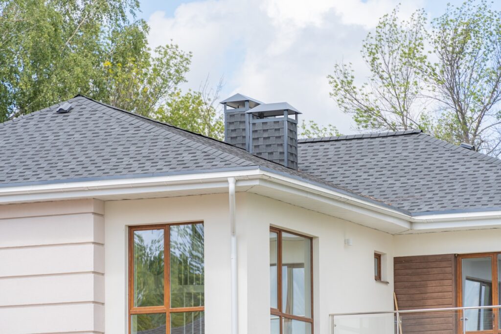 A modern house with light-colored walls, large windows, and a gray shingled roof by Roofing Ruskin featuring two chimney vents. Green trees and a blue sky with clouds are in the background.