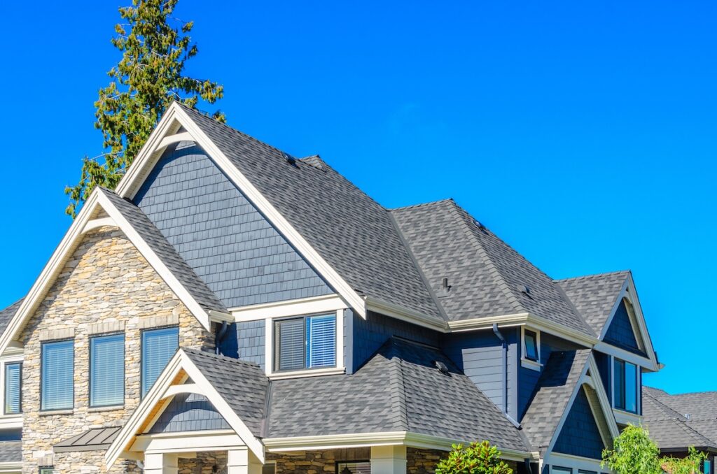 Close-up view of the upper part of a modern house with dark gray shingles by roofing Largo, stone accents, blue siding, and white trim, set against a clear blue sky with a tall tree in the background.