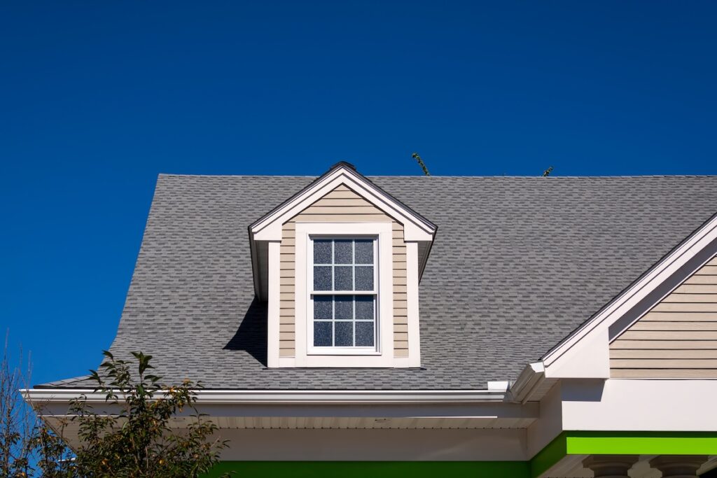 The photo shows the upper part of a house with gray shingles, a dormer window, beige siding, white trim, and a bright blue sky—an inviting example of quality roofing Land O Lakes homeowners can rely on.