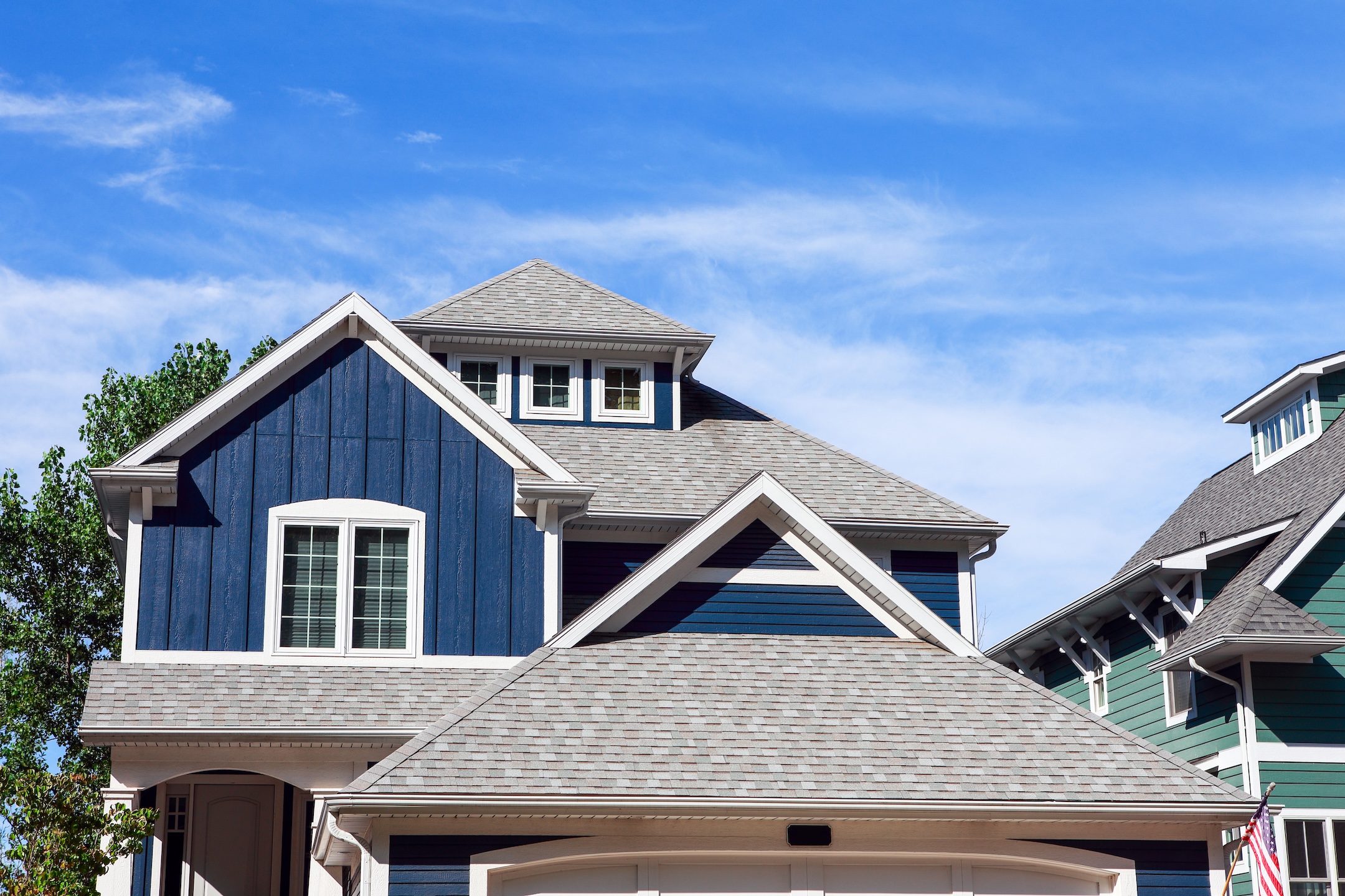 A modern house with blue siding, white trim, and gray shingles showcases expert roofing palm harbor craftsmanship, set against a bright blue sky with wispy clouds. Part of another green house is visible on the right.