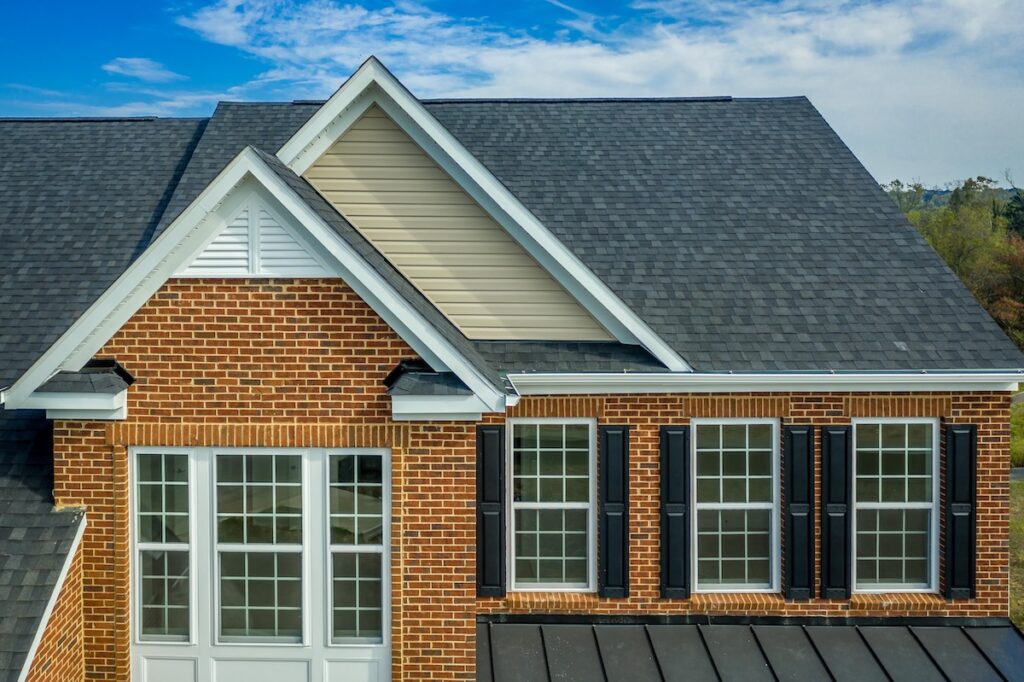 Close-up view of a house exterior in Palm Harbor featuring red brick walls, multiple white-trimmed windows with black shutters, and a dark gray shingled roof by roofing Palm Harbor experts under a partly cloudy sky.