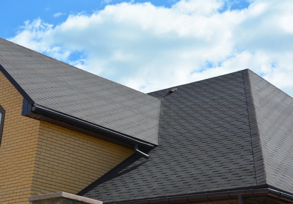Close-up of a house roof with gray asphalt shingles installed by roofing Largo experts, brown brick walls, and dark trim, set against a bright blue sky with scattered clouds.