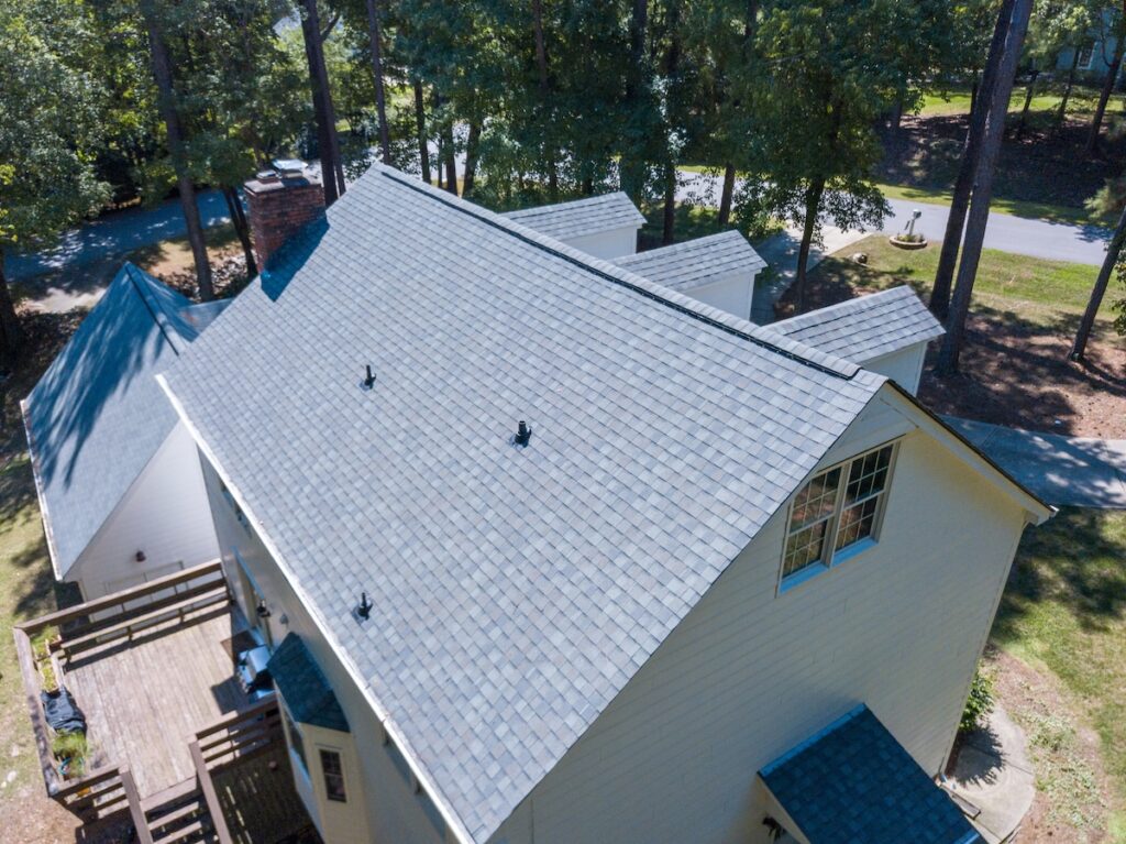 Aerial view of a light-colored house with a gray shingled roof by roofing St Petersburg, surrounded by trees. The roof features multiple vents and dormers, and a wooden deck is visible on the left side of the house.