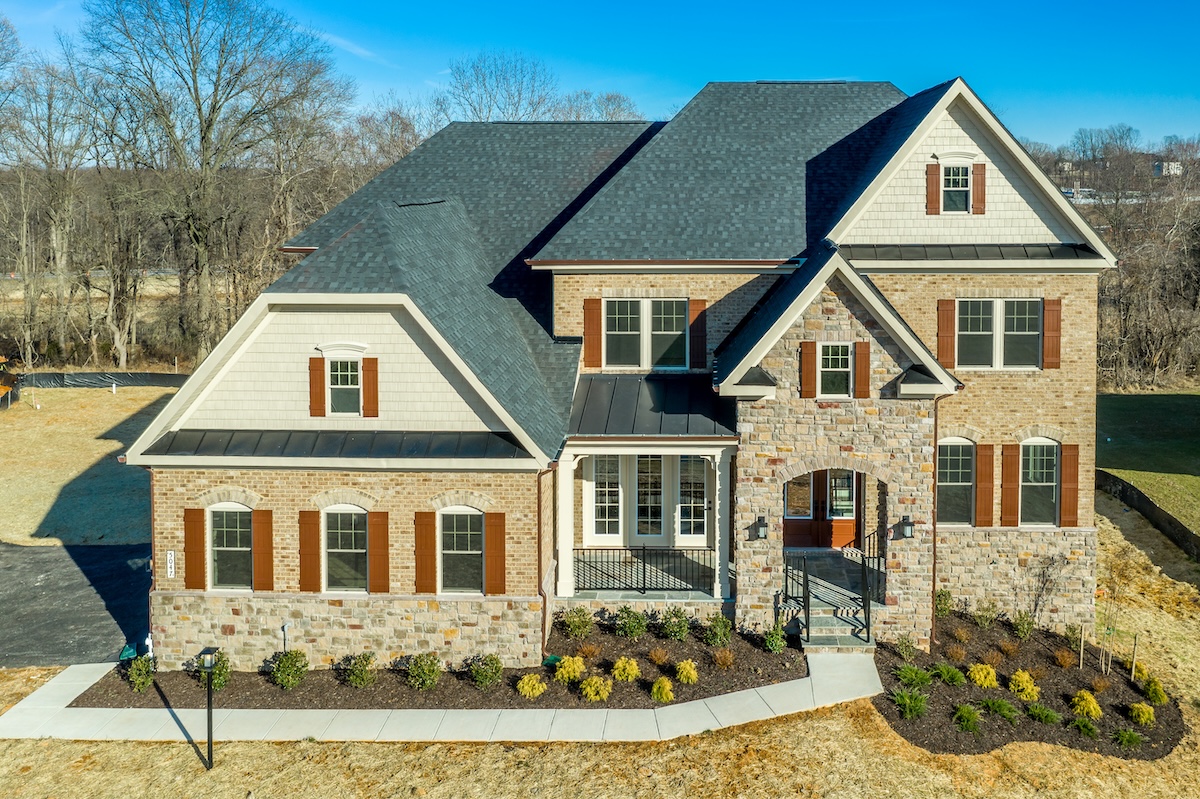A large two-story house with stone and brick exterior, multiple windows with brown shutters, new roofing Largo, a covered front porch, and landscaped yard on a clear, sunny day.