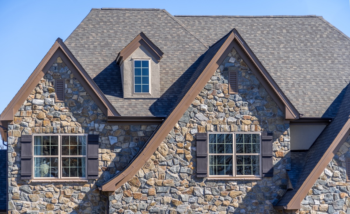 A close-up view of a house exterior featuring stone walls, three large windows with shutters, and a steep, multi-peaked shingle roof installed by Roofing Ruskin under a clear blue sky.