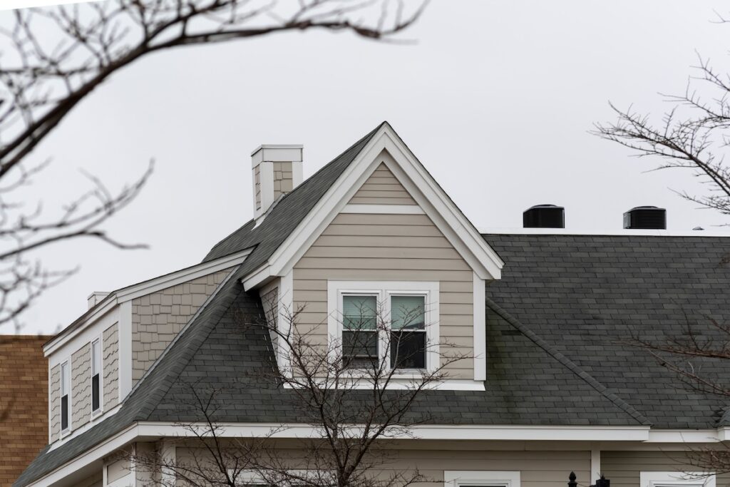 A close-up view of the upper part of a house with gray shingles and beige siding highlights expert roofing Citrus Park style, with a peaked roof partially framed by bare tree branches on a cloudy day.