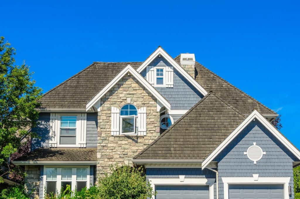 Two-story suburban house with gray shingles featuring expert roofing Tampa style, stone accents, white window shutters, and a double garage. Green shrubs and trees partially frame the home against a clear blue sky.