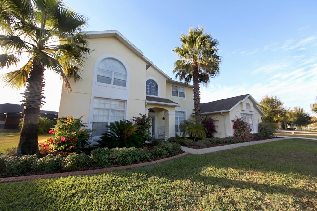 A two-story cream-colored house with large arched windows, surrounded by palm trees, shrubs, and flowering plants, sits under a blue sky with a well-maintained lawn—showcasing expert roofing Tampa craftsmanship.