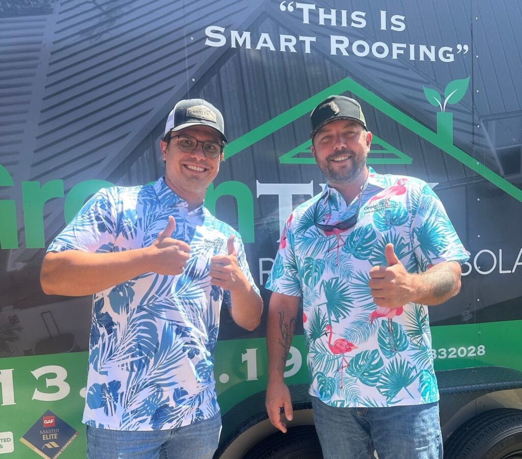 Two men wearing tropical-patterned shirts and baseball caps stand smiling in front of a vehicle with roofing advertisement graphics. Both are giving thumbs up and appear cheerful and relaxed.