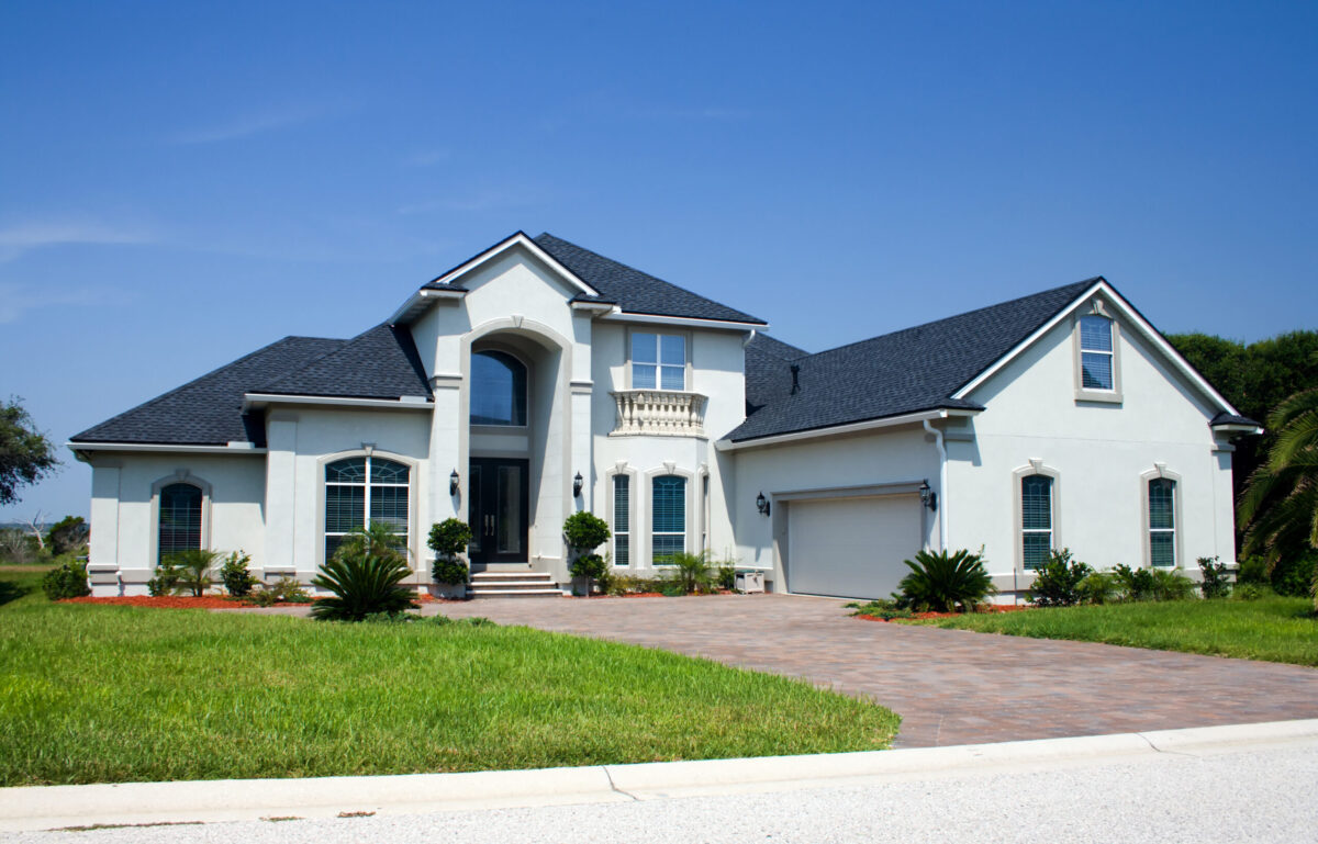 A large, modern two-story house with light-colored exterior walls, arched windows, a double-door entrance with columns, a dark shingled roof crafted by roofing Tampa experts, a three-car garage, and a manicured lawn under a blue sky.
