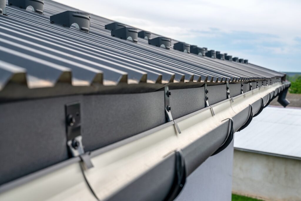 Close-up view of a modern metal roof with black ridges and sleek gutters attached to the edge of a building, set against a bright, slightly cloudy sky.