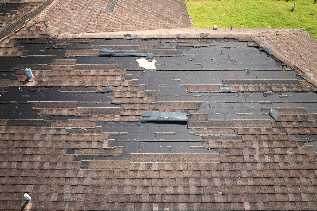 A roof with many missing and damaged brown shingles, revealing black underlayment underneath, shows clear signs of storm damage to roof. Debris is scattered across the worn shingles, with grass visible at the top right corner.
