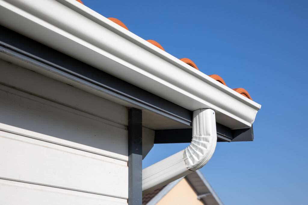 Close-up of white gutters and downspout attached to the edge of a house roof with red tiles, set against a clear blue sky.