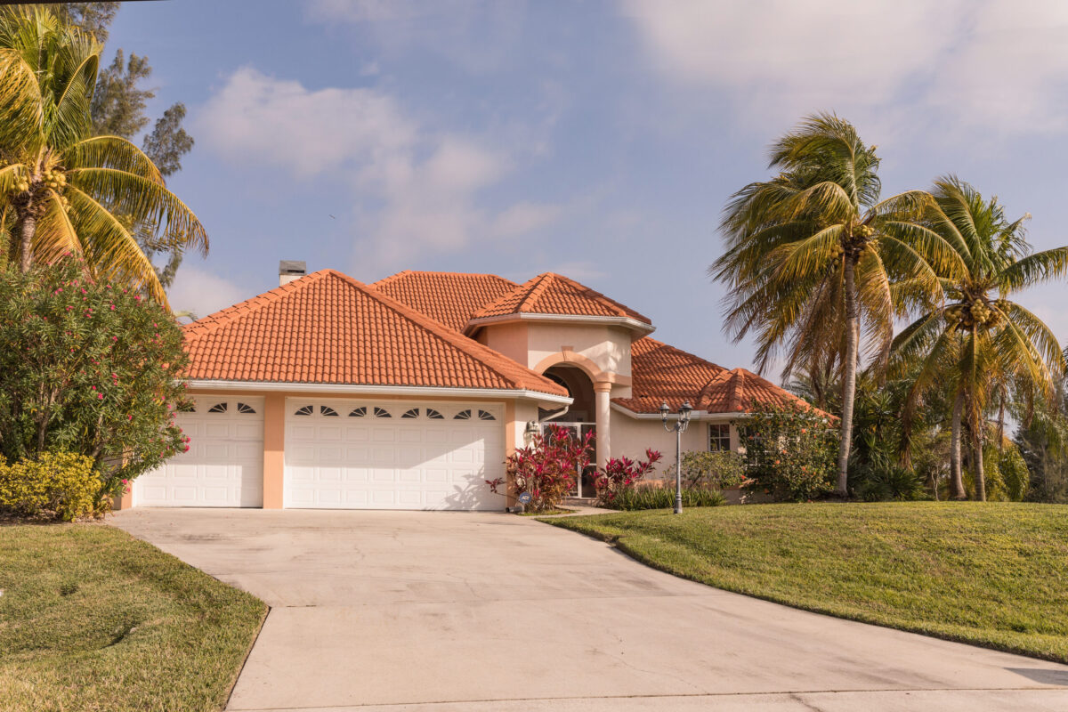 A single-story house with a red-tiled roof, well-maintained gutters, and a three-car garage, surrounded by palm trees and green lawn, under a partly cloudy sky.