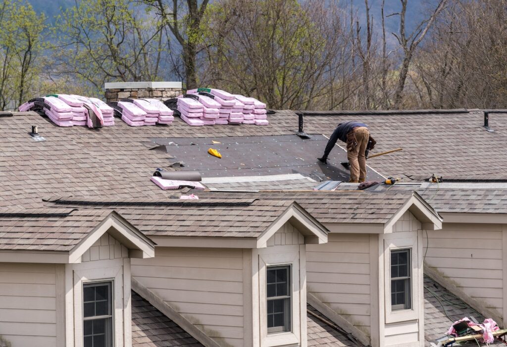 A worker repairs a shingle roof, surrounded by bundles of roofing materials, on a light-colored house with multiple dormer windows. Trees with early spring foliage are visible in the background. should i stay home during a roof replacement