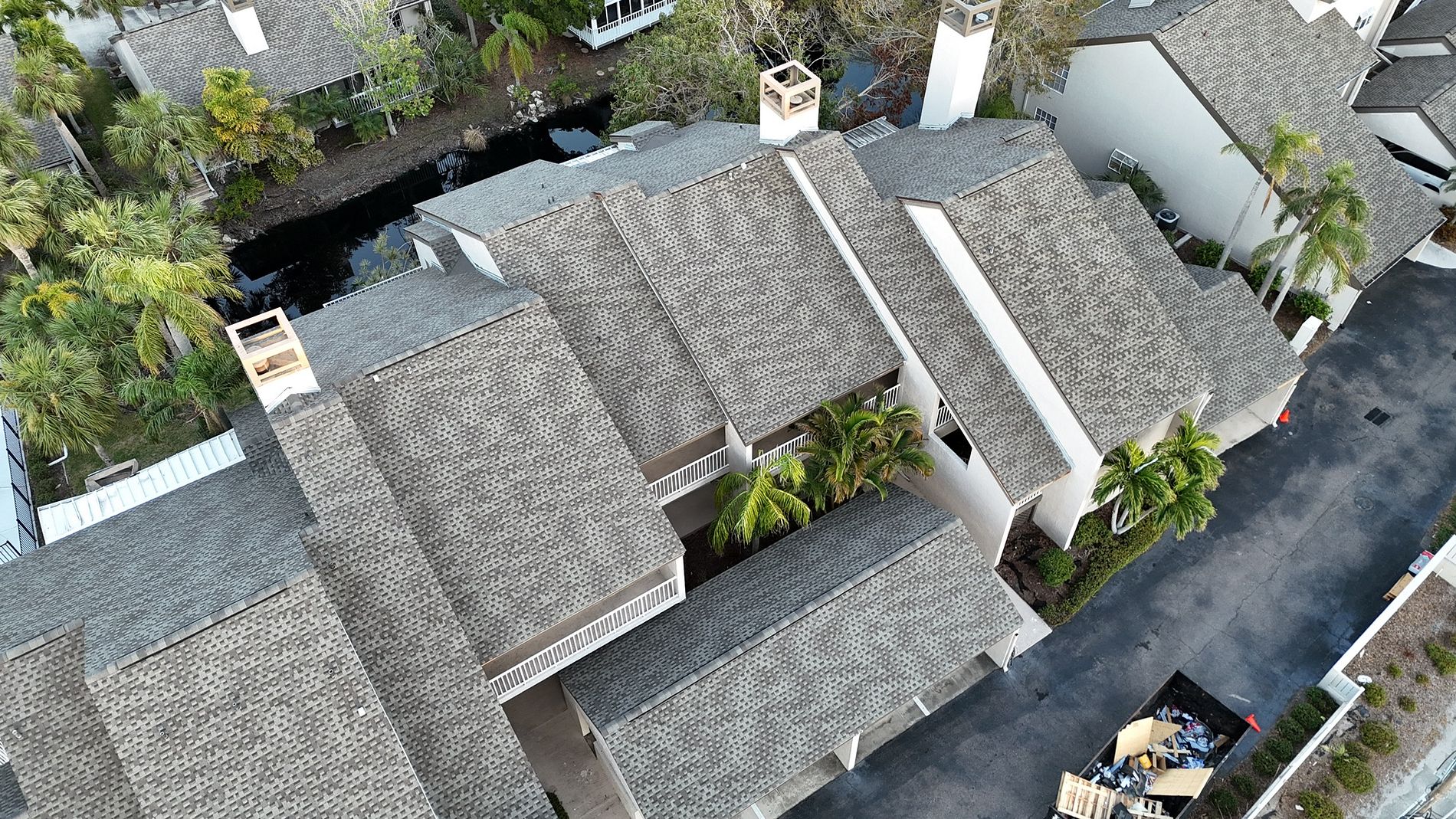 Aerial view of several modern houses with gray shingle roofs, surrounded by palm trees and a narrow road, with construction materials in one corner and a small waterway nearby.