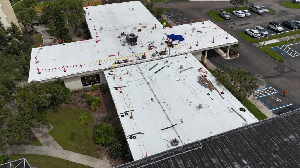 Aerial view of a building with a white flat roof under construction, scattered tools and materials, safety cones and flags, and nearby parked cars and trees.